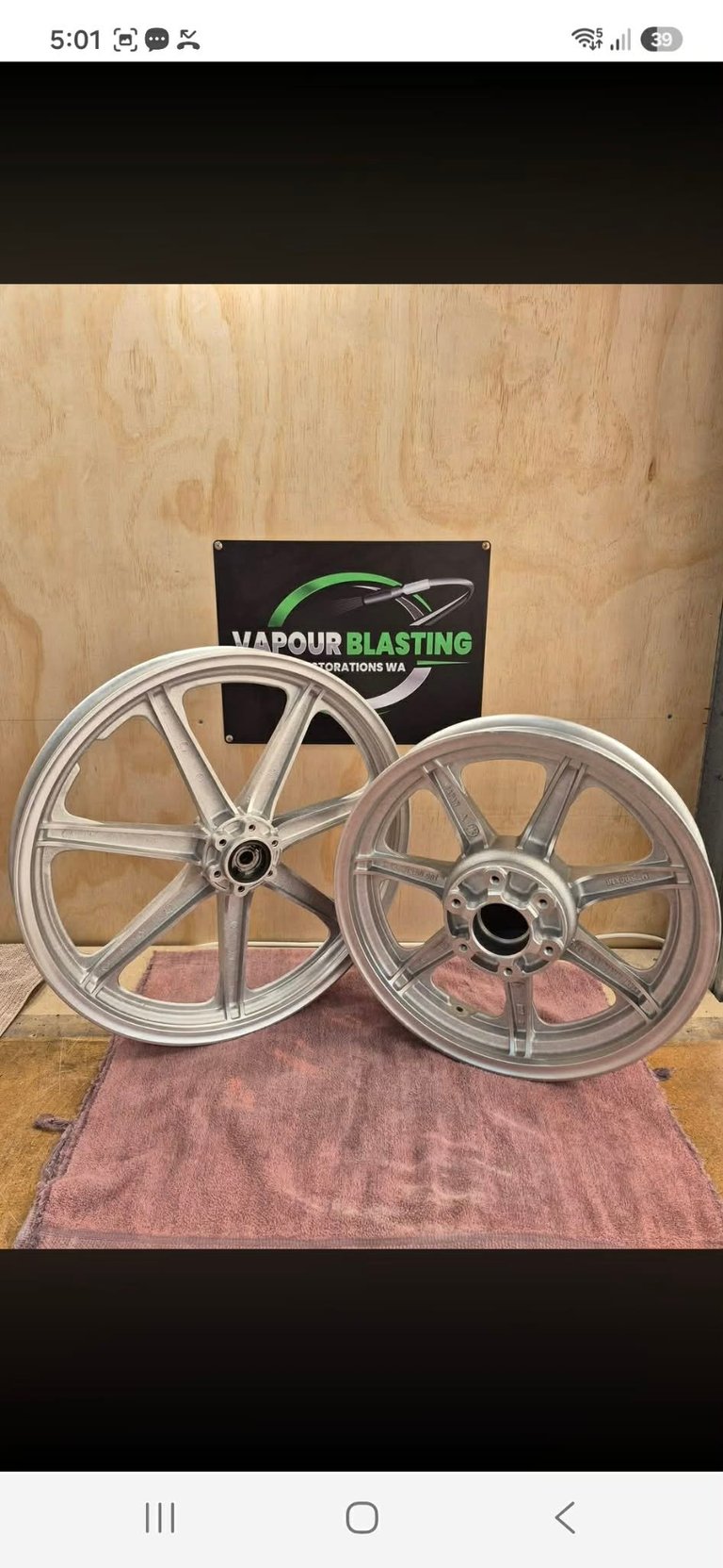 Two polished aluminum motorcycle wheels displayed on a pink mat against a plywood backdrop with a Vapour Blasting logo sign